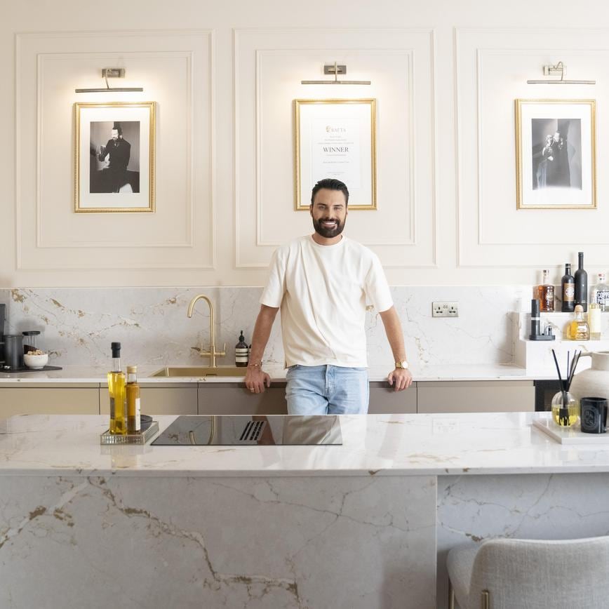 Rylan Clark stood in a modern kitchen with matt metallic bronze door fronts. It has an island with matching marble-effect worktops
